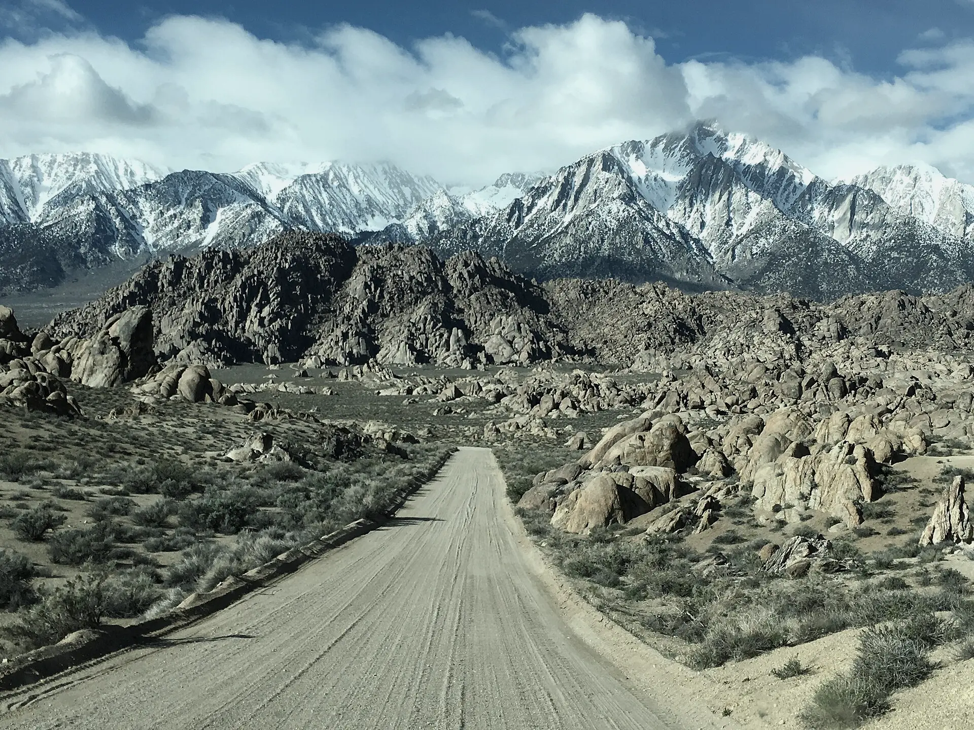 A graded dirt road leading through granite boulders toward snow-covered peaks under drifting clouds.