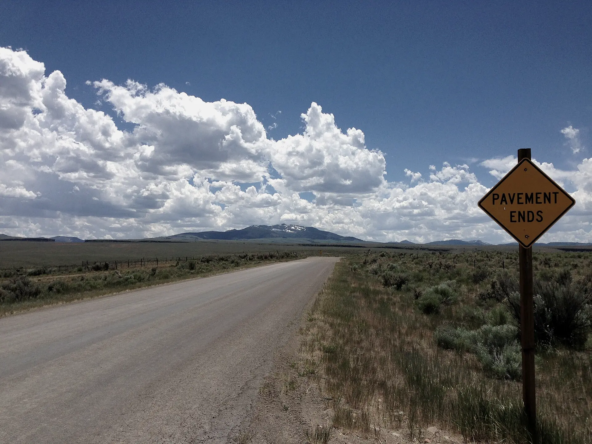 A “Pavement Ends” road sign beside a rural road that runs across sagebrush flats toward distant mountains.