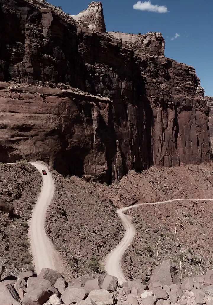 A dirt road switchbacking down beneath towering red sandstone cliffs.