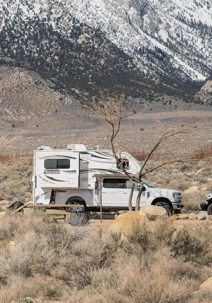 A pickup with a slide-in truck camper parked among desert scrub and boulders below a mountain slope.